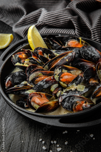 Baked mussels in spicy cheese sauce with lemon in a black cast-iron pot on wooden boards. Restaurant menu. Background black boards.