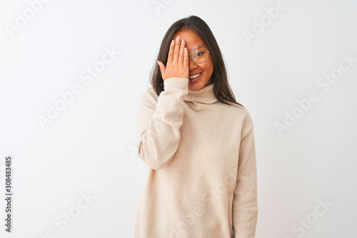 Young chinese woman wearing turtleneck sweater and glasses over isolated white background covering one eye with hand, confident smile on face and surprise emotion. © Krakenimages.com