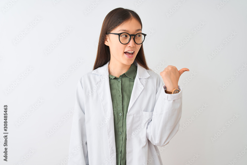 Young chinese scientist woman wearing coat and glasses over isolated white background smiling with happy face looking and pointing to the side with thumb up.