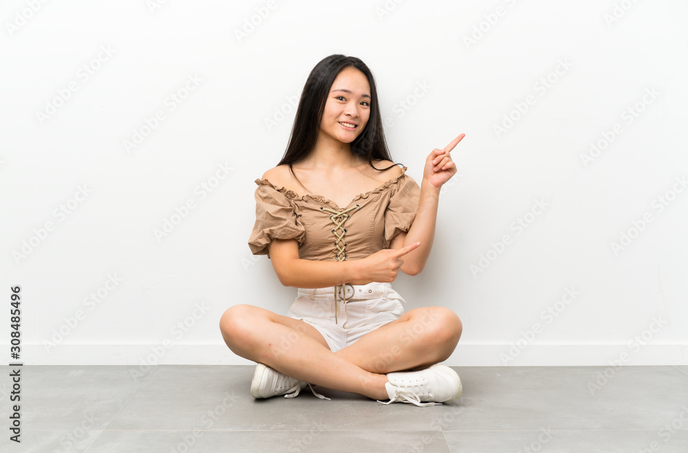 Teenager asian girl sitting on the floor pointing finger to the side Teenager asian girl sitting on the floor pointing finger to the side