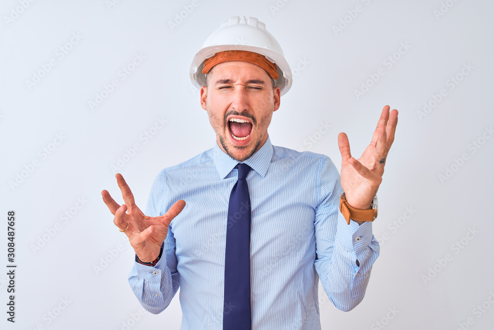 Young business man wearing contractor safety helmet over isolated background crazy and mad shouting and yelling with aggressive expression and arms raised. Frustration concept. Young business man wearing contractor safety helmet over isolated background crazy and mad shouting and yelling with aggressive expression and arms raised. Frustration concept.