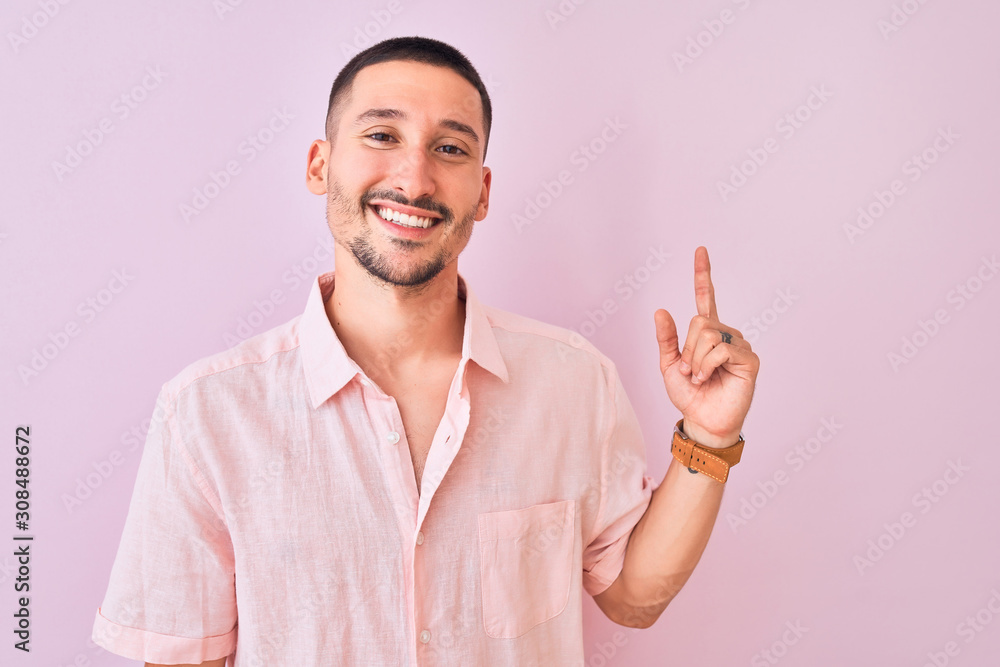 Young handsome man wearing pink shirt standing over isolated background with a big smile on face, pointing with hand and finger to the side looking at the camera. Young handsome man wearing pink shirt standing over isolated background with a big smile on face, pointing with hand and finger to the side looking at the camera.