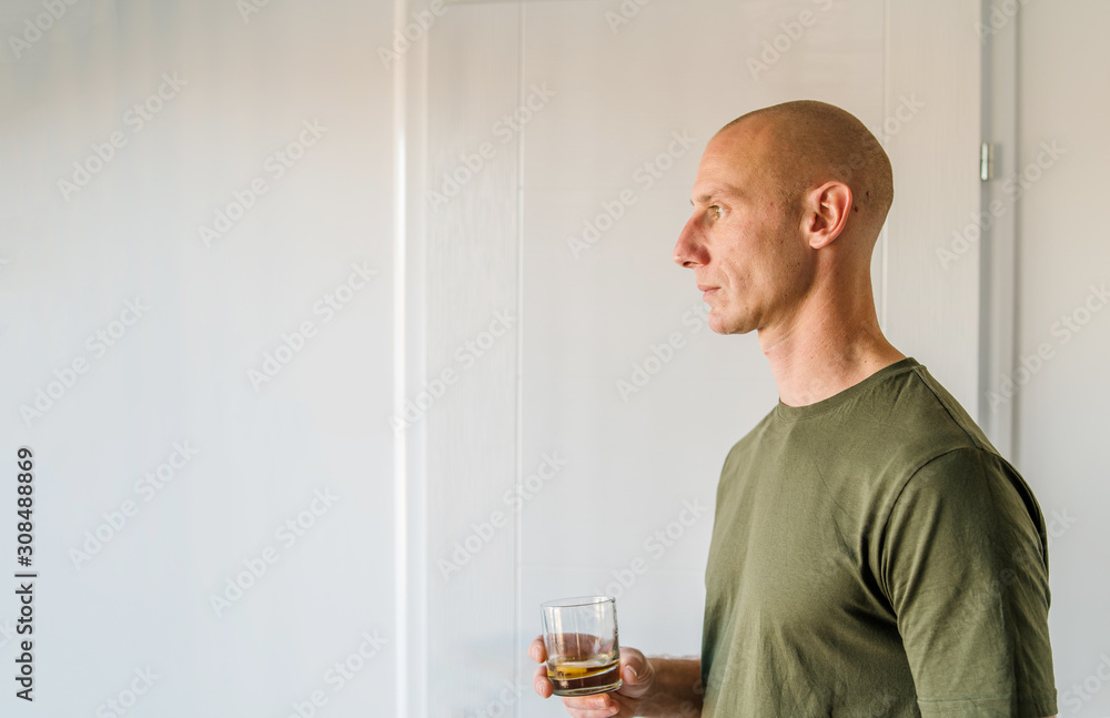 Portrait of young caucasian man good looking with short hair wearing green t shirt holding a glass of brandy or whiskey alcohol drink standing in front of white wall at home Portrait of young caucasian man good looking with short hair wearing green t shirt holding a glass of brandy or whiskey alcohol drink standing in front of white wall at home