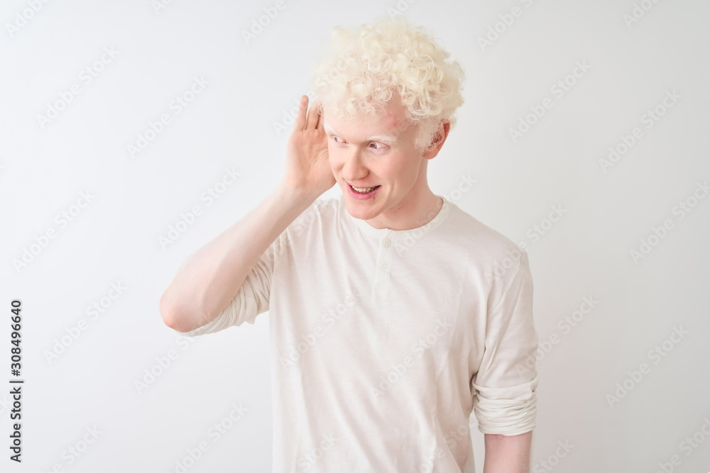 Young albino blond man wearing casual t-shirt standing over isolated white background smiling with hand over ear listening an hearing to rumor or gossip. Deafness concept. Young albino blond man wearing casual t-shirt standing over isolated white background smiling with hand over ear listening an hearing to rumor or gossip. Deafness concept.