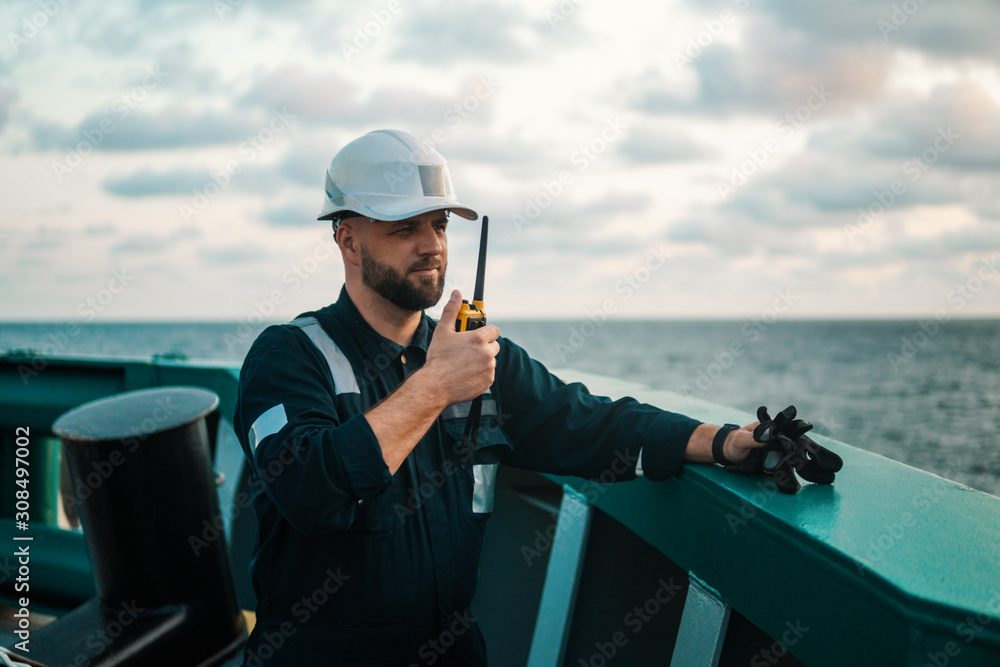 Obraz premium Marine Deck Officer or Chief mate on deck of offshore vessel or ship , wearing PPE personal protective equipment - helmet, coverall. He holds VHF walkie-talkie radio in hands.