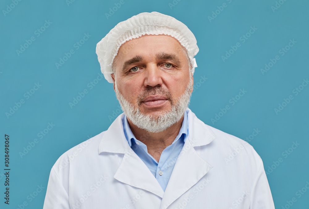 Portrait of mature surgeon in cap and in white coat looking at camera over blue background Portrait of mature surgeon in cap and in white coat looking at camera over blue background