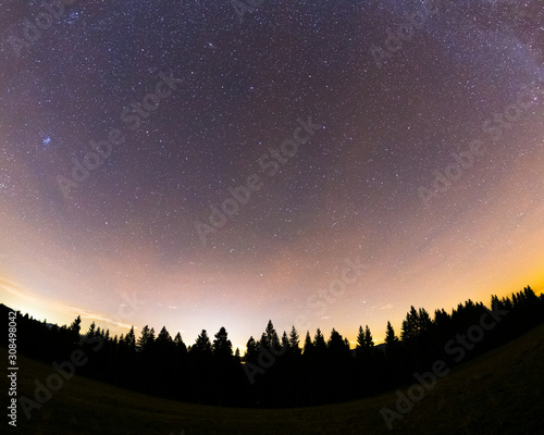 The Milky Way captured wide-spread along with the treetops with a yellow-orange glow on the horizon and lots of stars in the sky during a full night.