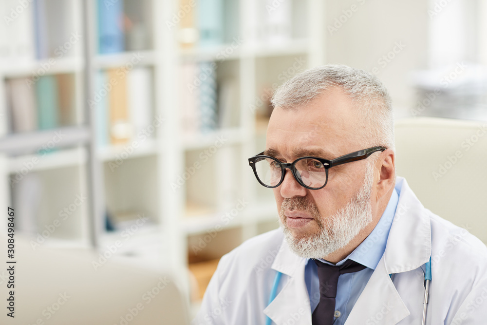 Mature doctor in eyeglasses looking at computer monitor and concentrating on his work Mature doctor in eyeglasses looking at computer monitor and concentrating on his work