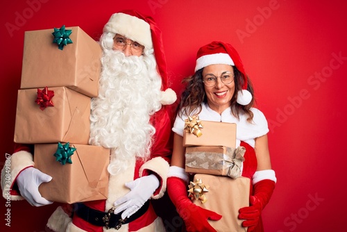 Middle age couple wearing Santa costume holding tower of gifts over isolated red background with a happy and cool smile on face. Lucky person.