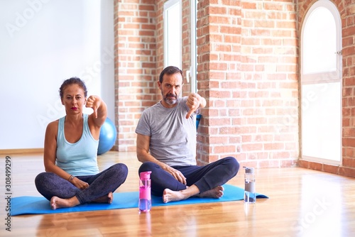 Middle age sporty couple sitting on mat doing stretching yoga exercise at gym looking unhappy and angry showing rejection and negative with thumbs down gesture. Bad expression.