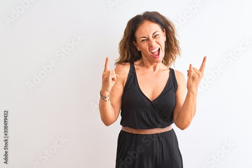 Middle age woman wearing black casual dress standing over isolated white background shouting with crazy expression doing rock symbol with hands up. Music star. Heavy concept.