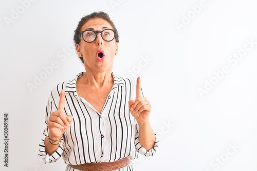 Quadro su tela Middle age businesswoman wearing striped dress and glasses over isolated white background amazed and surprised looking up and pointing with fingers and raised arms