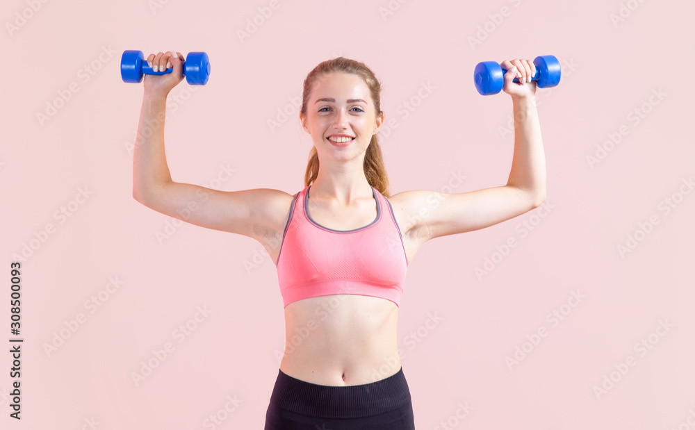 Young woman with two dumbbells in hands, standing on a pink background. Fitness exercise.