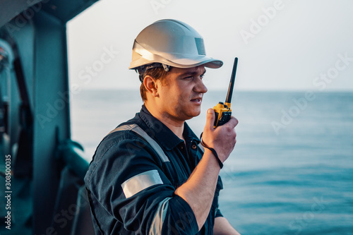 Marine Deck Officer or Chief mate on deck of offshore vessel or ship , wearing PPE personal protective equipment - helmet, coverall. He holds VHF walkie-talkie radio in hands.