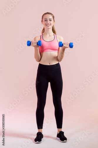 Young woman with two dumbbells in hands, standing on a pink background. Fitness exercise.