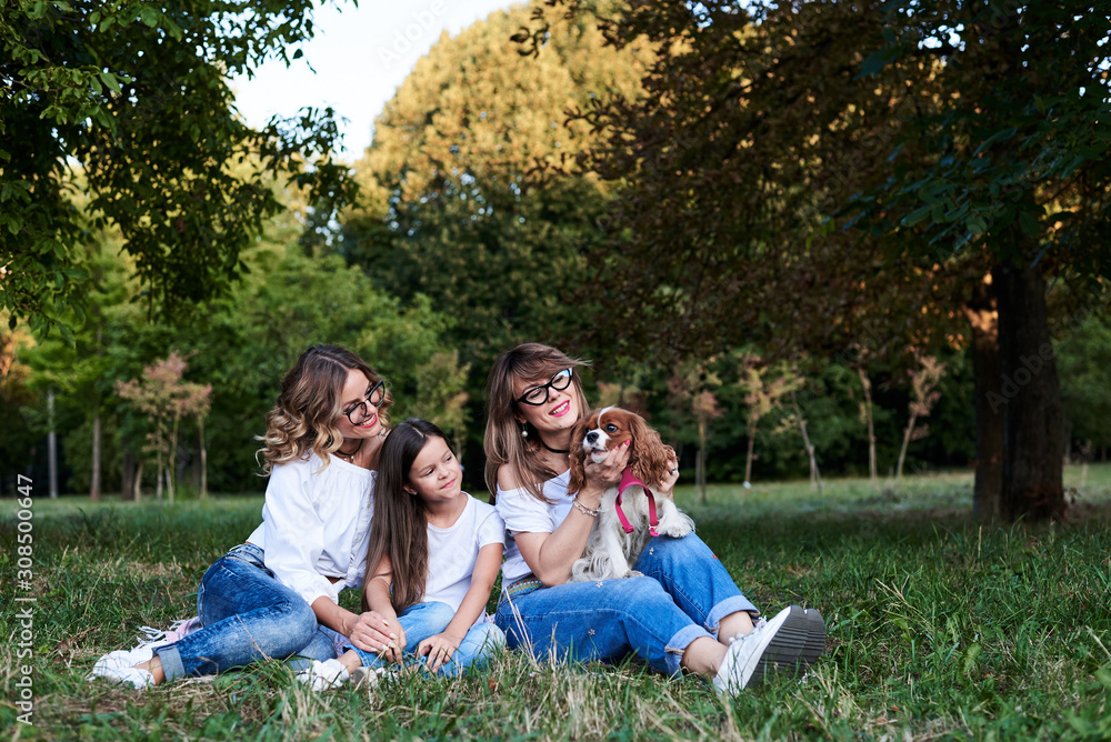 Two young blond women and one brunette girl, wearing jeans and white t-shirts, sitting on green grass in park, playing with cavalier king charles spaniel, smiling, laughing. Leisure time in summer.