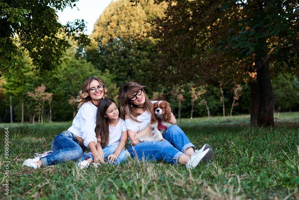 Two young blond women and one brunette girl, wearing jeans and white t-shirts, sitting on green grass in park, playing with cavalier king charles spaniel, smiling, laughing. Leisure time in summer. Two young blond women and one brunette girl, wearing jeans and white t-shirts, sitting on green grass in park, playing with cavalier king charles spaniel, smiling, laughing. Leisure time in summer.