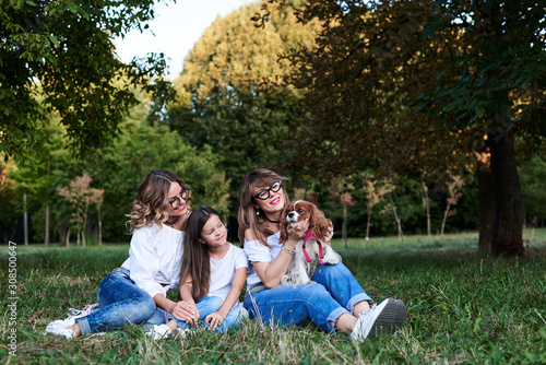 Two young blond women and one brunette girl  wearing jeans and white t-shirts  sitting on green grass in park  playing with cavalier king charles spaniel  smiling  laughing. Leisure time in summer.