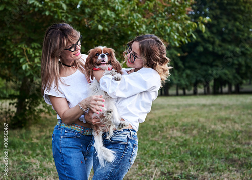 Two young blond women, wearing white t-shirts,, holding small white and brown dog in park in summer, smiling. Cavalier king charles spaniel with his owners on a walk.