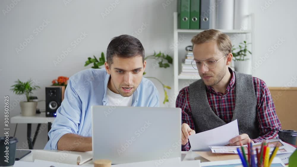Zoom in medium shot of two creative young designers examining and discussing patterns on laptop and samples printed on paper sitting at desk in office Zoom in medium shot of two creative young designers examining and discussing patterns on laptop and samples printed on paper sitting at desk in office