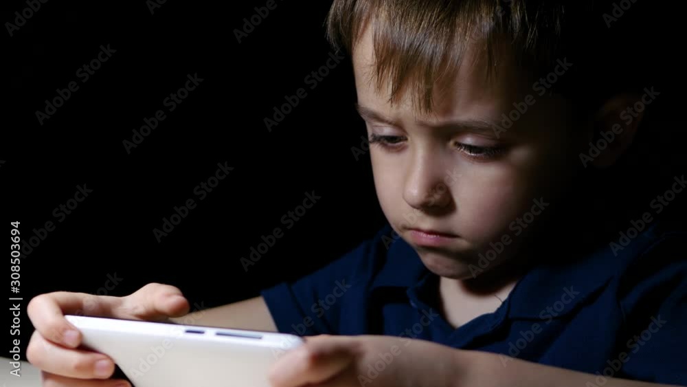 A little boy spends time watching a movie on his smartphone. The child plays on the phone in a dark room. A little boy spends time watching a movie on his smartphone. The child plays on the phone in a dark room.
