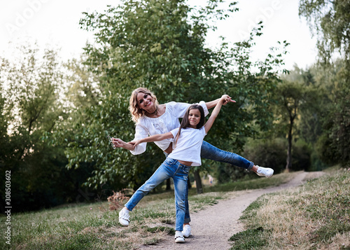 Young blond mother posing for picture with her brunette daughter in the park in summer. Woman and girl, wearing blue jeans and white t-shirts, having fun, laughing, smiling. Family leisure fun time.