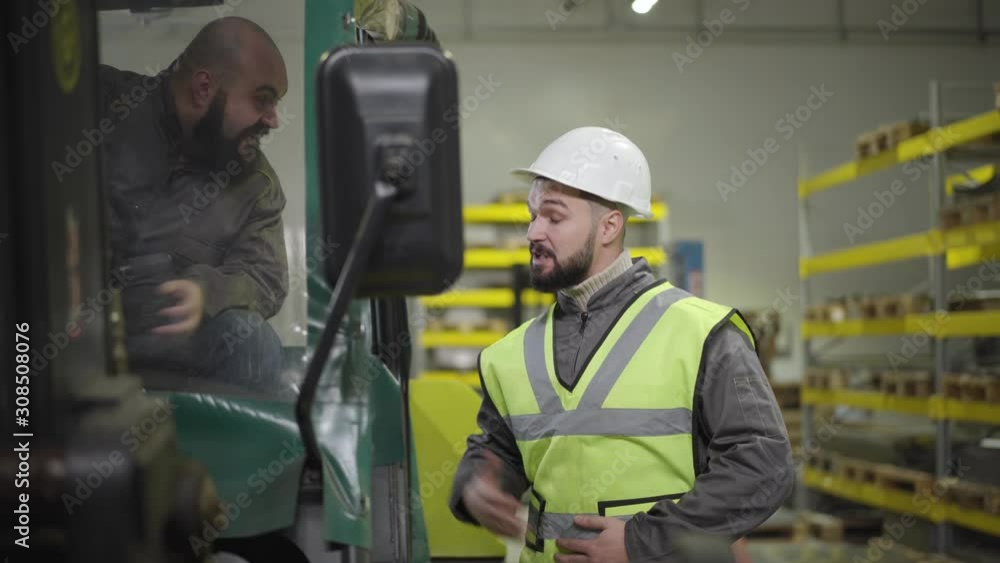 Portrait of tow tractor driver talking to Caucasian man in helmet and vest. Workers having break at production site. Manufacture, working, warehouse, industry. Portrait of tow tractor driver talking to Caucasian man in helmet and vest. Workers having break at production site. Manufacture, working, warehouse, industry.