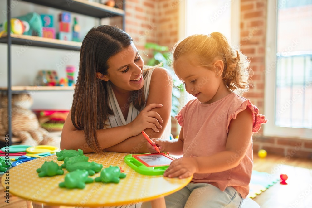 Fototapeta premium Beautiful teacher and blond student toddler girl drawing using digital board at kindergarten