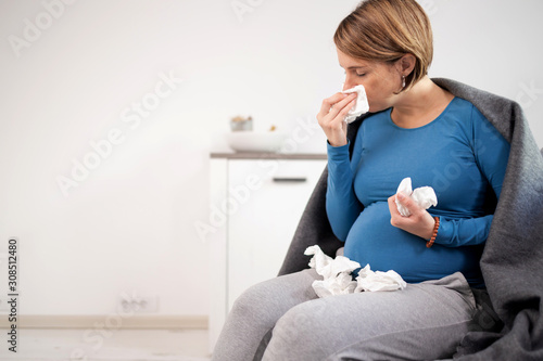 Pregnant woman catching cold, flu, virus, sitting at home on a couch, blowing nose and beaing sick. photo