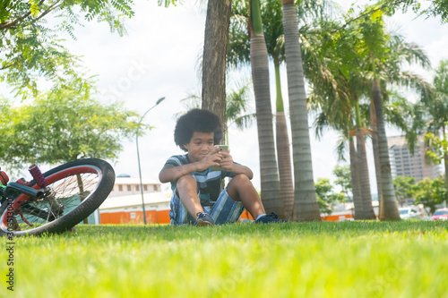 The young boy sits on the grass in the garden and using smartphone. A bicycle lying on the grass under the tree.