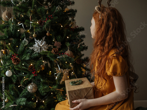 Girl holding a Christmas gift on her lap on the background of the Christmas tree