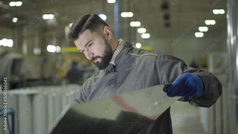 Bearded Caucasian man in working gloves standing at steel metal pipes holding metal sheet. Young worker checking material for production of ready items. Steel plant, manufacturing, metal production. Bearded Caucasian man in working gloves standing at steel metal pipes holding metal sheet. Young worker checking material for production of ready items. Steel plant, manufacturing, metal production.