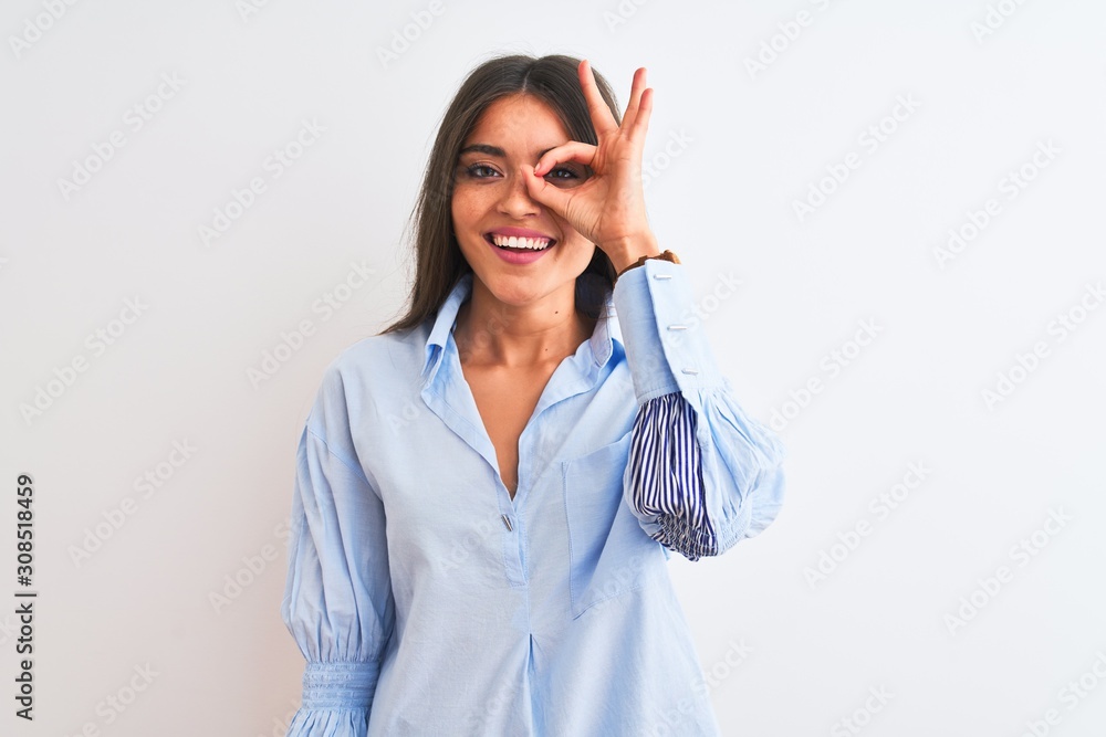 Young beautiful woman wearing blue elegant shirt standing over isolated white background doing ok gesture with hand smiling, eye looking through fingers with happy face. Young beautiful woman wearing blue elegant shirt standing over isolated white background doing ok gesture with hand smiling, eye looking through fingers with happy face.