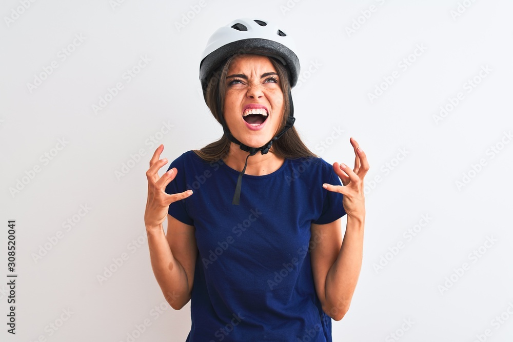 Young beautiful cyclist woman wearing security bike helmet over isolated white background crazy and mad shouting and yelling with aggressive expression and arms raised. Frustration concept.