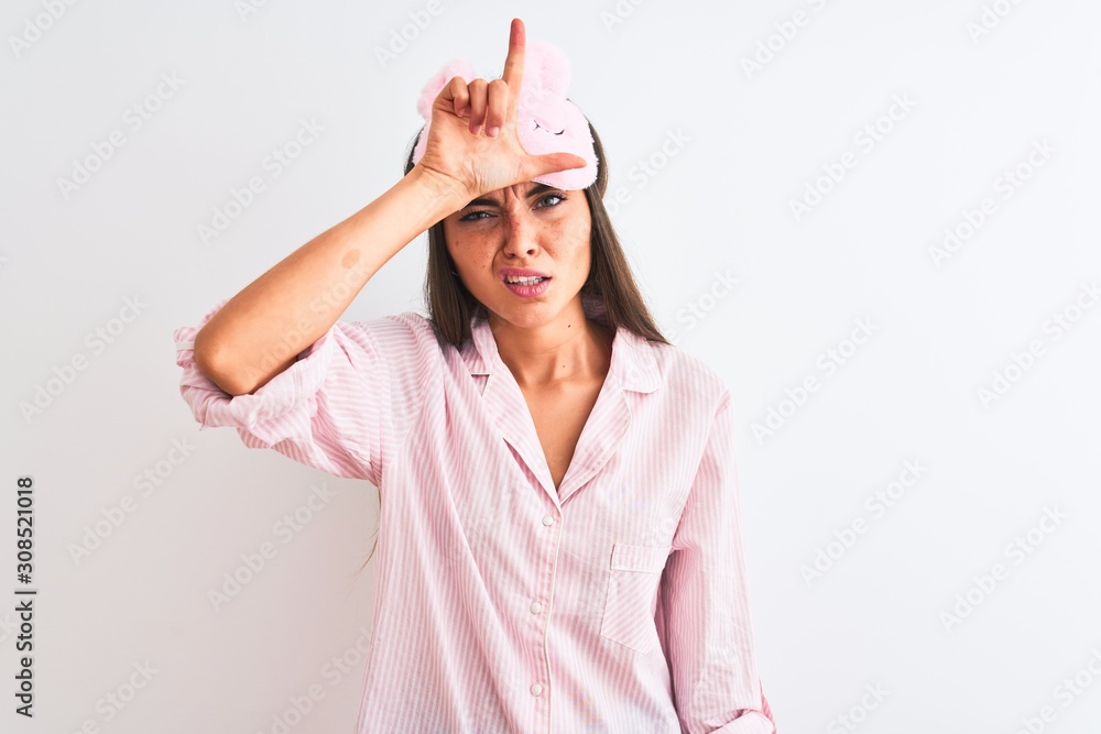 Young beautiful woman wearing sleep mask and pajama over isolated white background making fun of people with fingers on forehead doing loser gesture mocking and insulting. Young beautiful woman wearing sleep mask and pajama over isolated white background making fun of people with fingers on forehead doing loser gesture mocking and insulting.