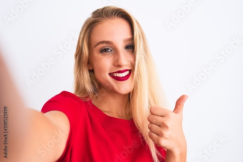 Young beautiful woman wearing t-shirt make selfie by camera over isolated white background happy with big smile doing ok sign, thumb up with fingers, excellent sign © Krakenimages.com