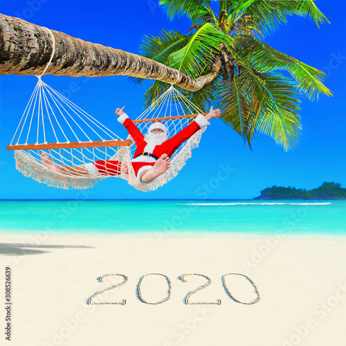Santa Claus thumbs up hands relax in mesh hammock under coconut palm tree at paradise ocean beach with caption happy New Year 2020 on sand, Christmas travel destination concept