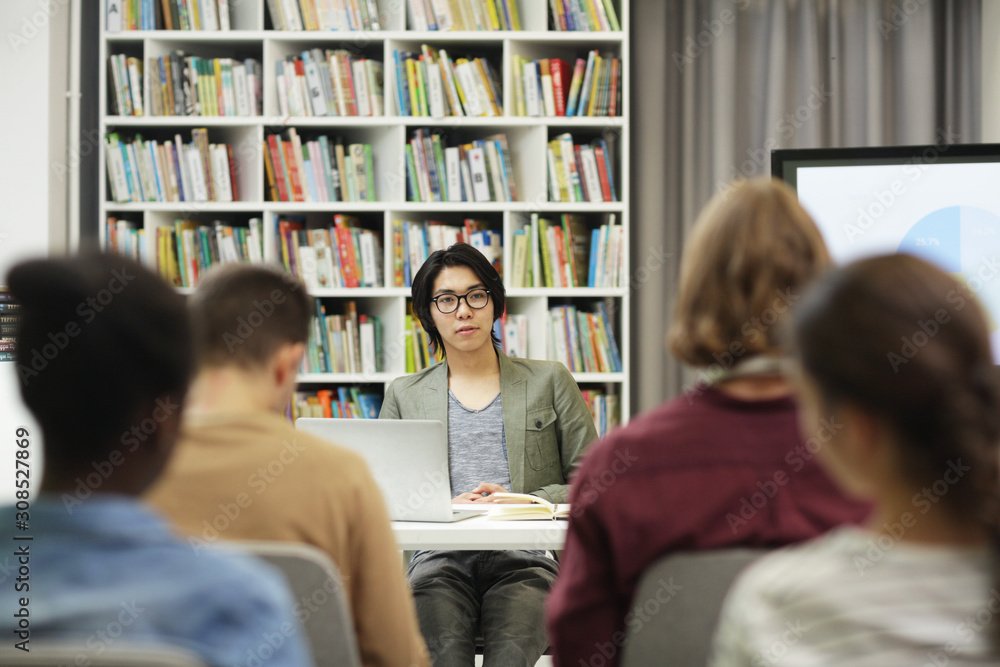 Young Asian writer sitting at the table and presenting his new book to the audience in the library Young Asian writer sitting at the table and presenting his new book to the audience in the library