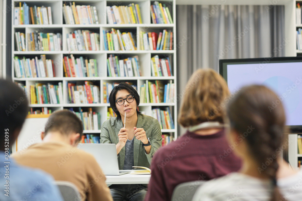 Asian young teacher in eyeglasses sitting at desk and explaining the new material to the group of students in the library Asian young teacher in eyeglasses sitting at desk and explaining the new material to the group of students in the library