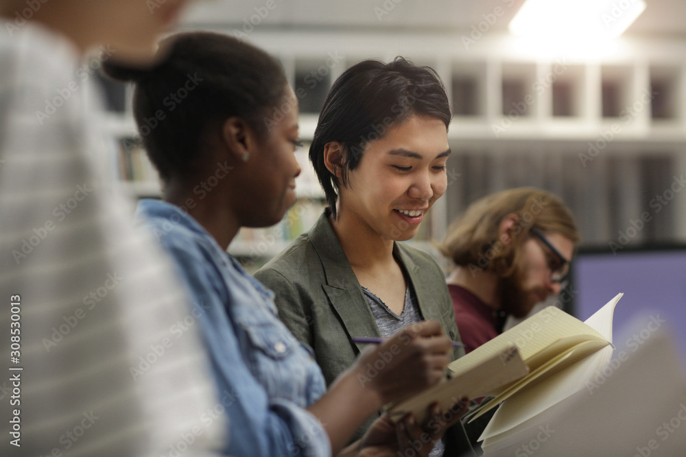 Two multiethnic students sitting together looking at their notebooks and discussing work together during lesson Two multiethnic students sitting together looking at their notebooks and discussing work together during lesson