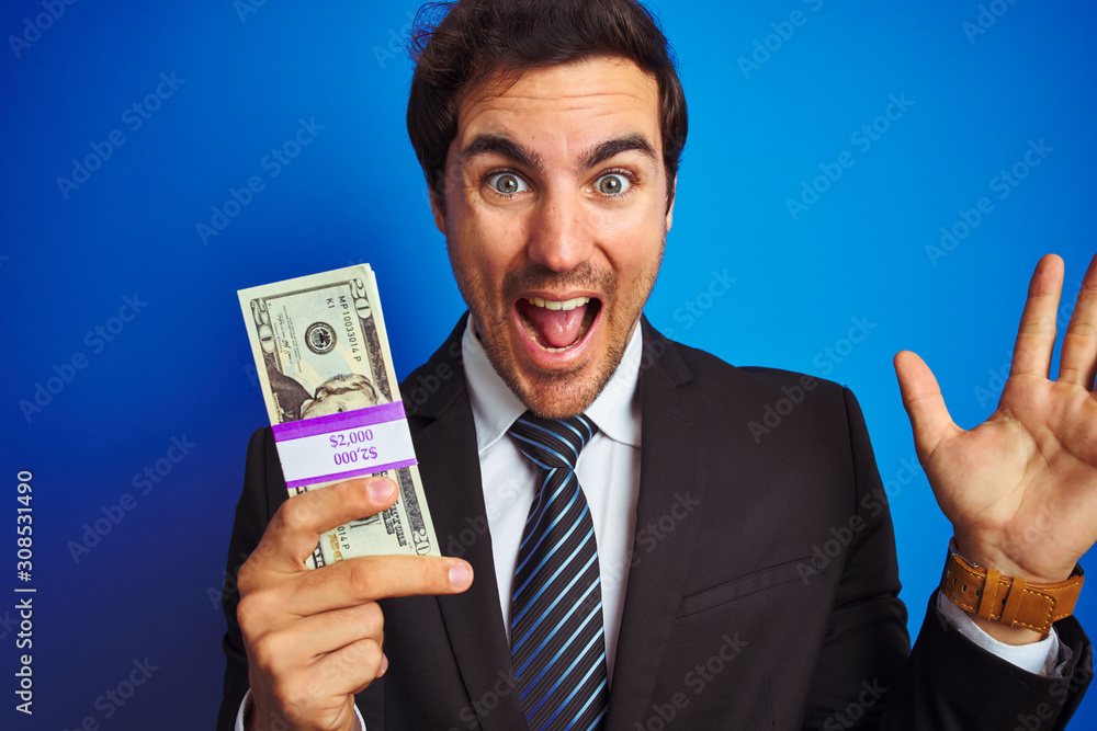 Young handsome businessman wearing suit holding dollars over isolated blue background very happy and excited, winner expression celebrating victory screaming with big smile and raised hands Young handsome businessman wearing suit holding dollars over isolated blue background very happy and excited, winner expression celebrating victory screaming with big smile and raised hands