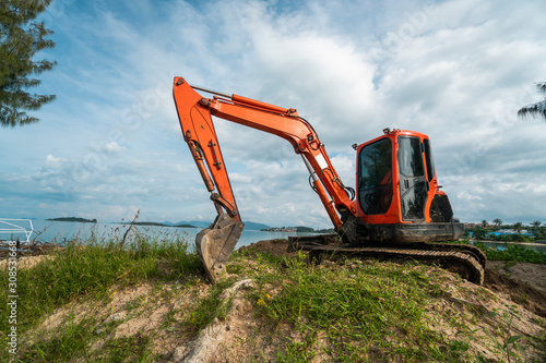 Small orange excavator on a ground against blue sky and sea for a works on construction site. Small tracked excavator standing on a ground with a blue sea on background. Heavy industry.