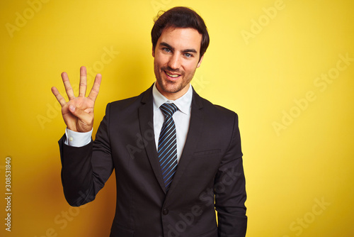 Young handsome businessman wearing suit and tie standing over isolated yellow background showing and pointing up with fingers number four while smiling confident and happy.