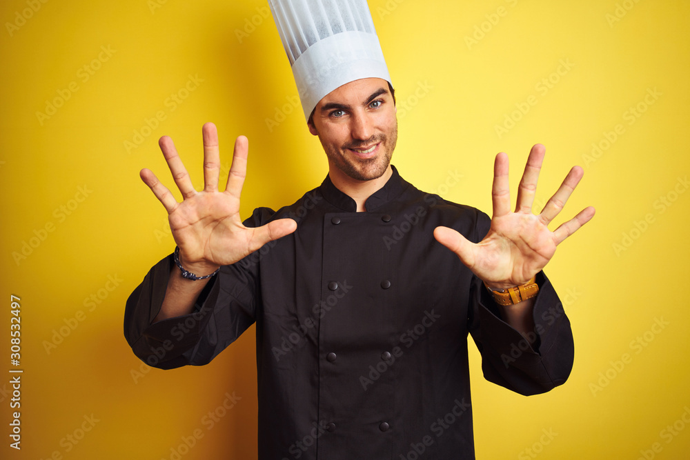 Young chef man wearing uniform and hat standing over isolated yellow background showing and pointing up with fingers number ten while smiling confident and happy.