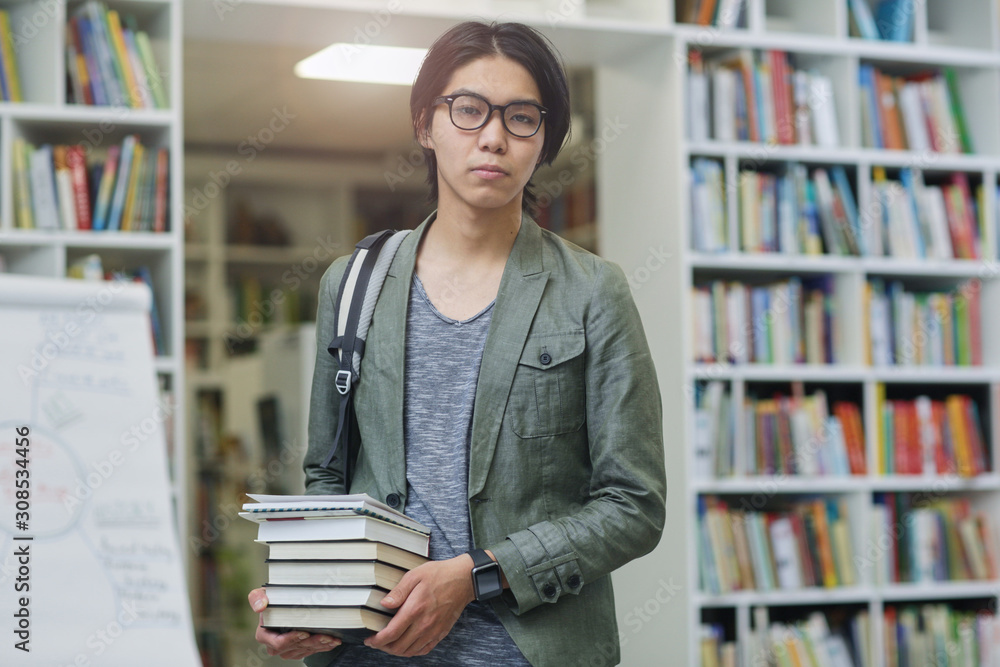 Portrait of Asian young student in eyeglasses holding heap of books and looking at camera he comes to the library