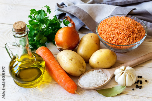 Ingredients for vegetarian soup puree with red lentils: few yellow potatoes, carrot, onion and garlic, olive oil, parsley. On a white textured wooden table.