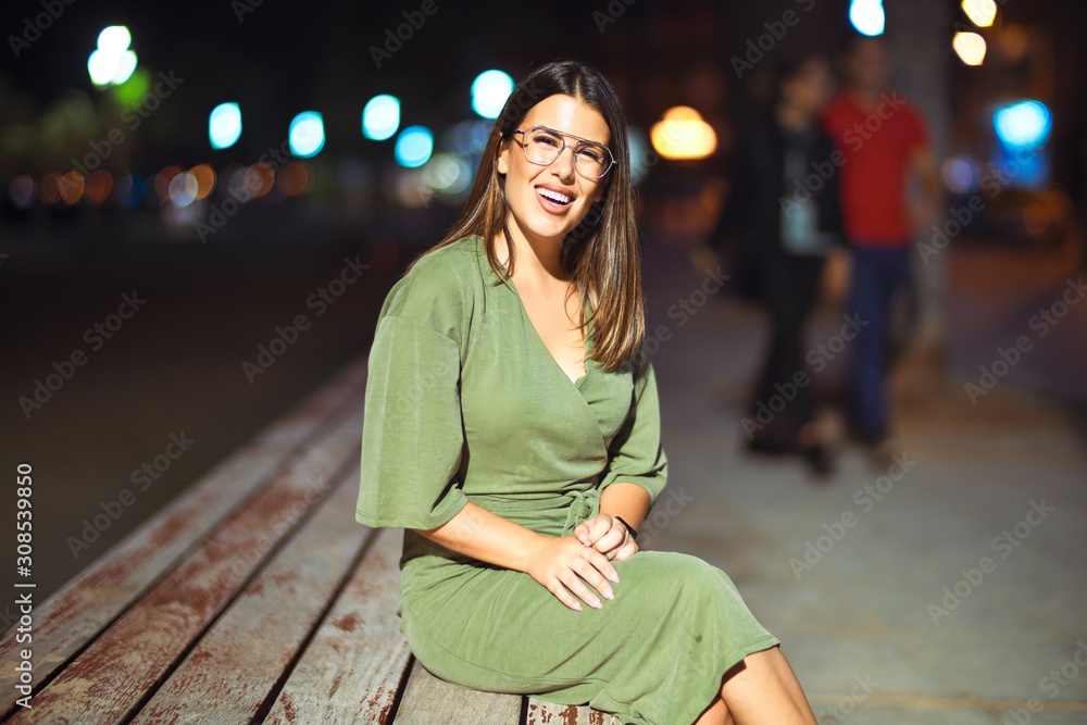 Fototapeta premium Young beautiful girl smiling happy and confident relaxing on bench at the promenade, sitting with a smile on face around night lights bokeh