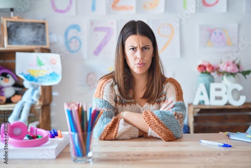 Young beautiful teacher woman wearing sweater and glasses sitting on desk at kindergarten skeptic and nervous, disapproving expression on face with crossed arms. Negative person.