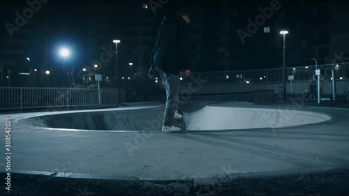 A young guy comes to the pool with a skateboard and prepares to jump into the pit. Skateboarder rides a skateboard at night on the street, lights are on. Teenager on a skateboard. Rear view, 4K