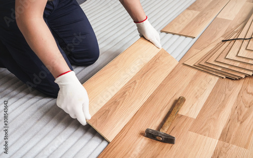 Installing laminated floor, detail on man hands in white gloves, holding wooden tile, over white foam base layer, small hammer near